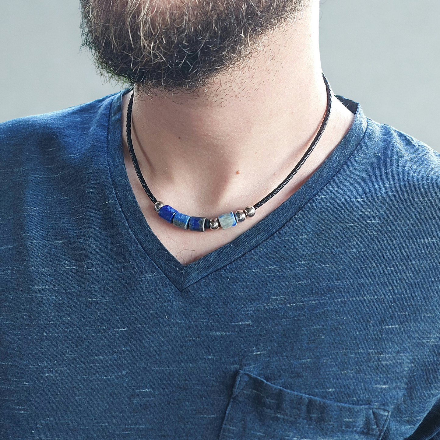 A close-up of a man's neck and chest wearing a black leather cord necklace with blue and staineless steel beads, paired with a blue V-neck t-shirt. The beads are a morse code message