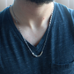 A close-up of a man's neck and chest wearing a morse code sterling silver chain necklace, paired with a dark blue v-neck t-shirt