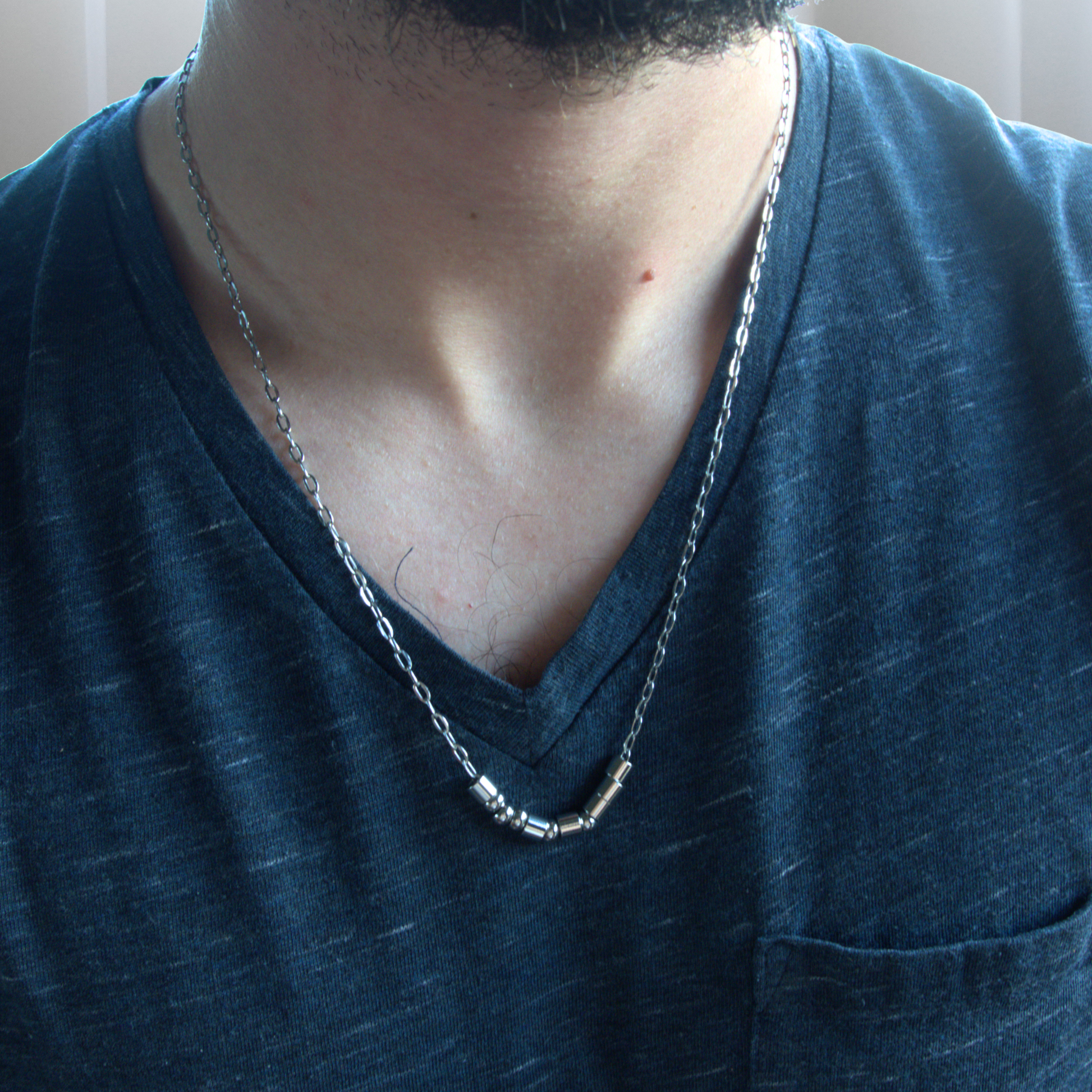 A close-up of a man's neck and chest wearing a morse code sterling silver chain necklace, paired with a dark blue v-neck t-shirt
