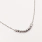 A close-up of a silver-toned chain necklace featuring a curved segment of polished metallic cylindrical beads and small spacer beads representing morse code, set against a plain off-white background.