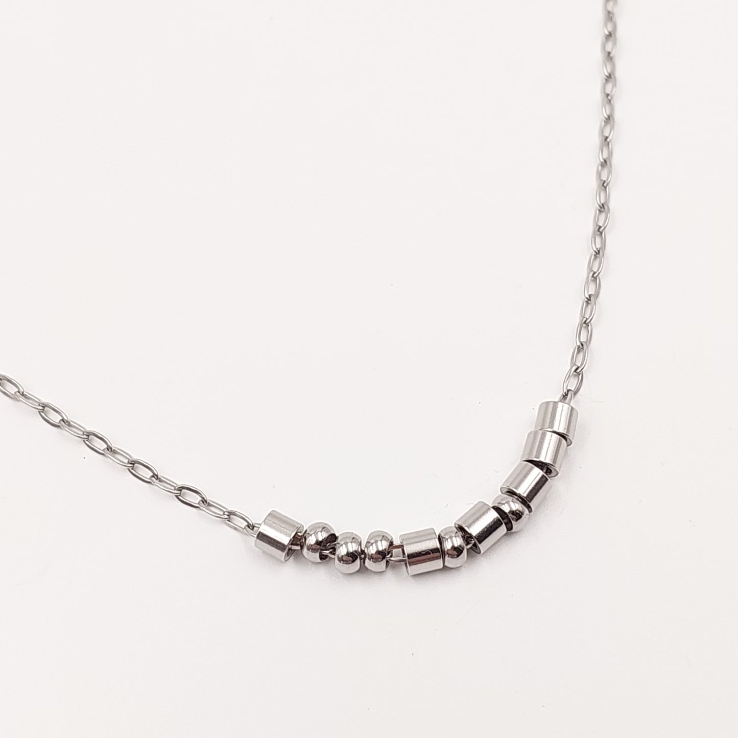 A close-up of a silver-toned chain necklace featuring a curved segment of polished metallic cylindrical beads and small spacer beads representing morse code, set against a plain off-white background.