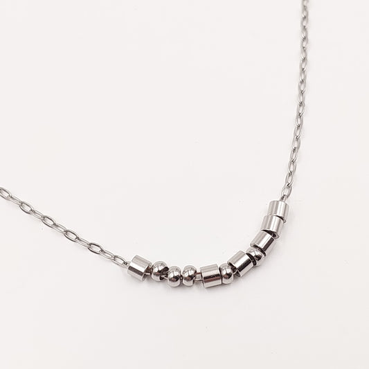A close-up of a silver-toned chain necklace featuring a curved segment of polished metallic cylindrical beads and small spacer beads representing morse code, set against a plain off-white background.